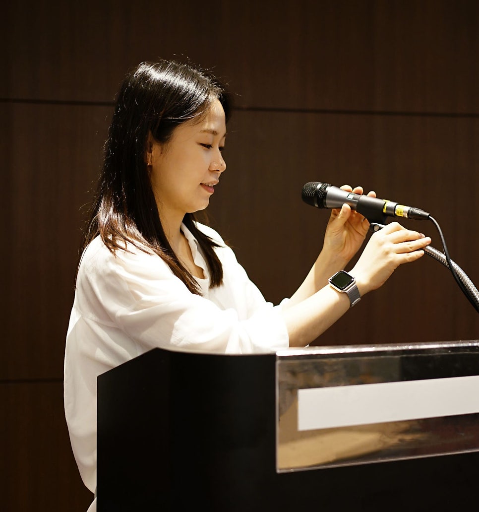 흰 셔츠를 입은 여성이 검은색 연단 뒤에서 마이크를 잡고 이야기하고 있다.