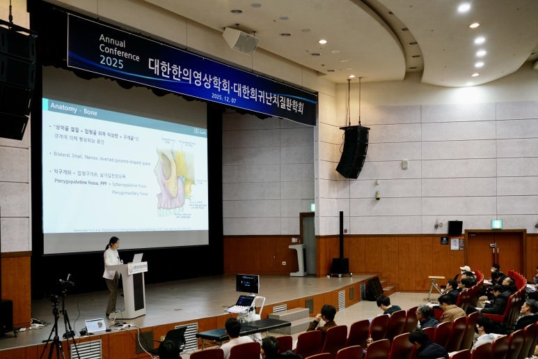 강당에서 한의학 학술대회가 열리고 있으며, 연단에 선 발표자가 해부학 관련 슬라이드를 보여주며 강연하고 있다. 무대에는 초음파 장비가 놓여 있고, 객석에는 청중들이 앉아 있다.