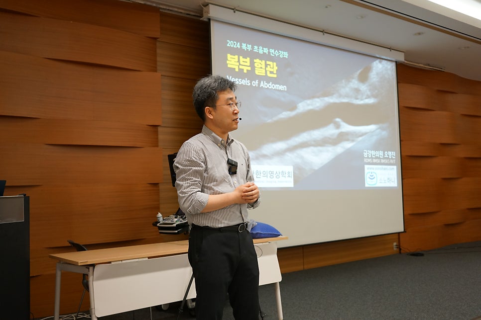 강사가 복부 초음파 영상이 보이는 화면 앞에서 발표하고 있는 모습
