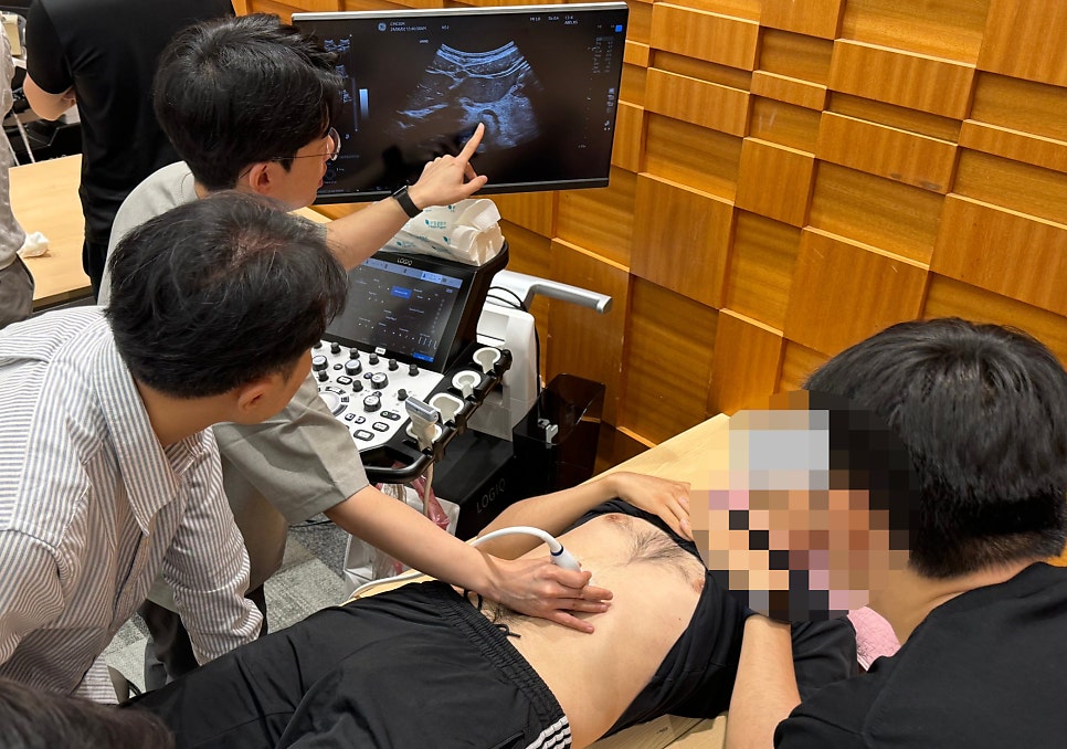 여러 사람이 초음파 기기 주변에 모여 있다. 한 사람이 누워있는 다른 사람의 복부에 초음파 탐촉자를 대고 있고, 또 다른 한 사람이 모니터에 표시된 초음파 영상을 가리키고 있다.