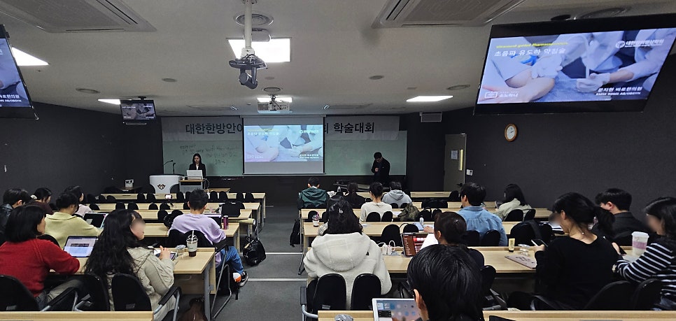 강의실에서 한의학 학술대회 발표자가 강단에 서 있고, 참석자들이 앉아 발표를 듣고 있다. 스크린에는 초음파 유도하 약침 관련 내용이 보인다.