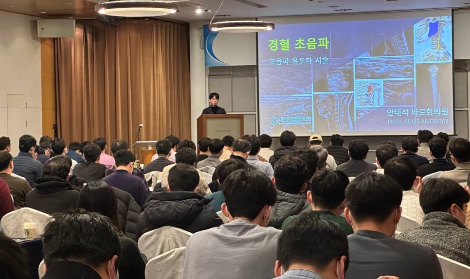 강사가 단상에서 경혈 초음파 관련 내용을 발표하고 있으며, 청중들이 앉아서 강연을 듣고 있는 모습.