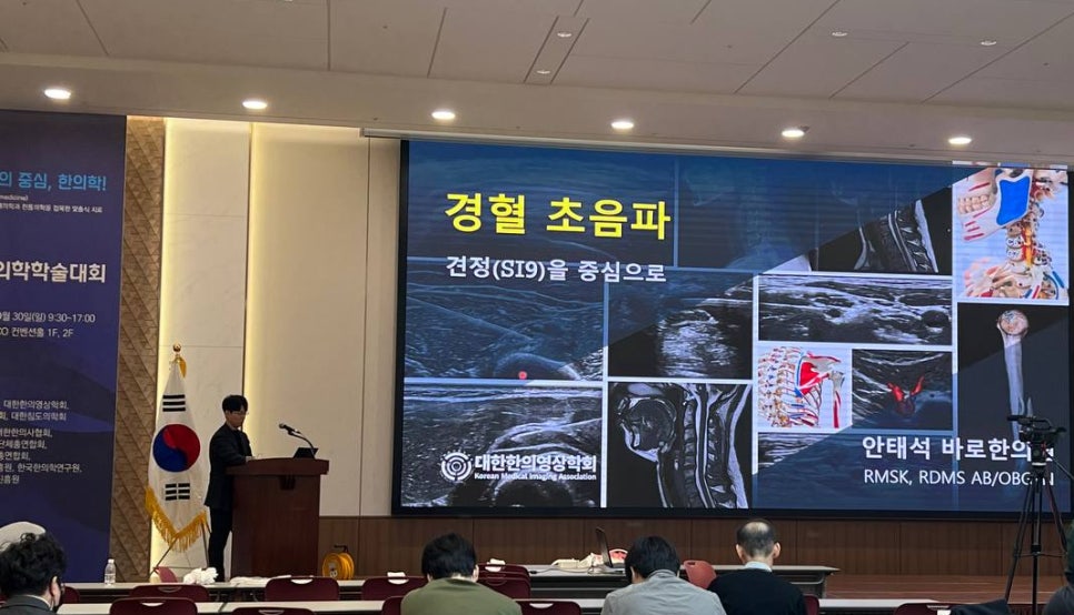 한의학 학술대회에서 강사가 경혈 초음파와 해부학 이미지가 있는 화면 앞에서 발표하는 모습 — 대한한의영상학회 초음파 약침·도침 강의 | 서울 바로한의원