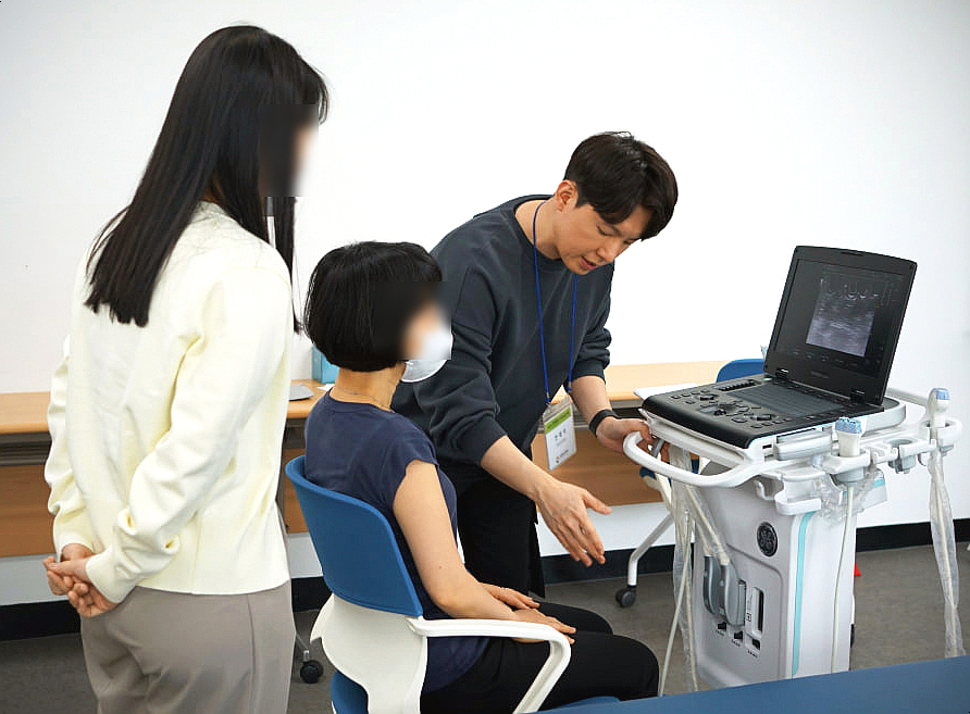 한 남성이 앉아있는 여성에게 초음파 진단기기 사용법을 설명하고 있으며, 다른 여성이 옆에서 교육을 참관하고 있다. 기기 화면에는 초음파 영상이 보인다.