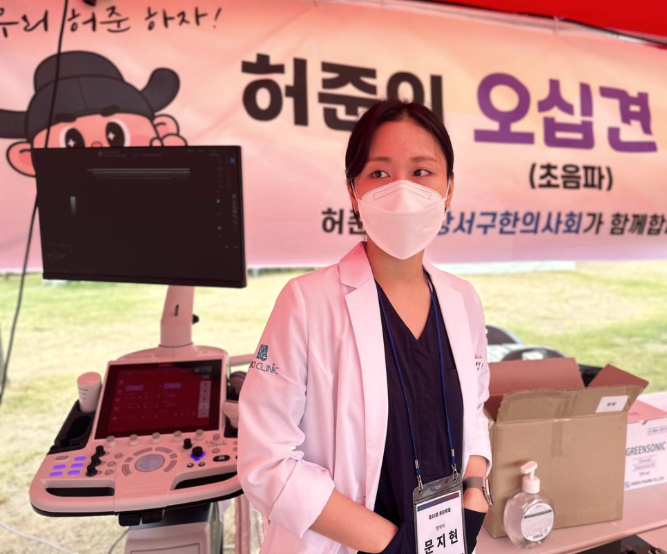 마스크를 쓴 여성이 흰색 가운을 입고 초음파 기기 옆에 서 있는 모습