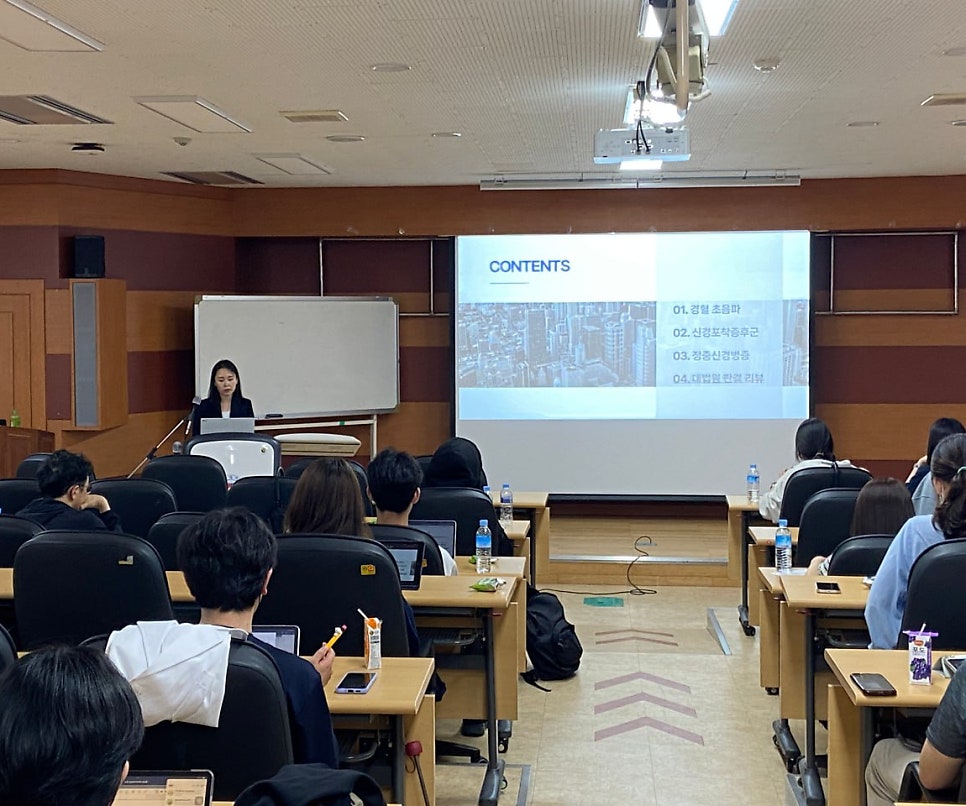 강의실에서 발표자가 강단에 서서 강연하고 있으며, 스크린에는 'CONTENTS'와 함께 '경혈 초음파' 등의 내용이 보인다. 여러 명의 청중이 앉아 발표를 듣고 있다.
