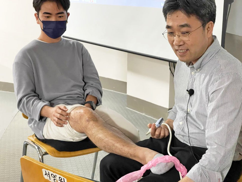 줄무늬 셔츠를 입은 남성이 마스크를 쓴 남성의 다리에 초음파 기기를 대고 시연하는 모습