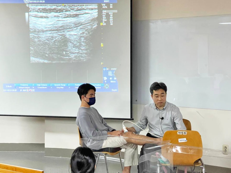 강사가 학생의 다리에 초음파 기기를 사용하여 시연하고 있으며, 스크린에 초음파 영상이 나타나 있다.