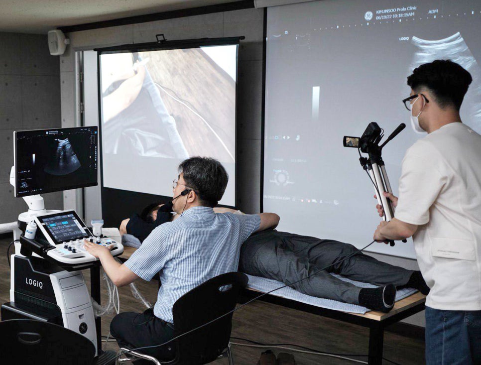 한의사가 엎드린 환자에게 초음파 검사를 하고 있으며, 초음파 영상이 모니터와 스크린에 표시되고 있다. 한 사람이 이 과정을 촬영하고 있다.