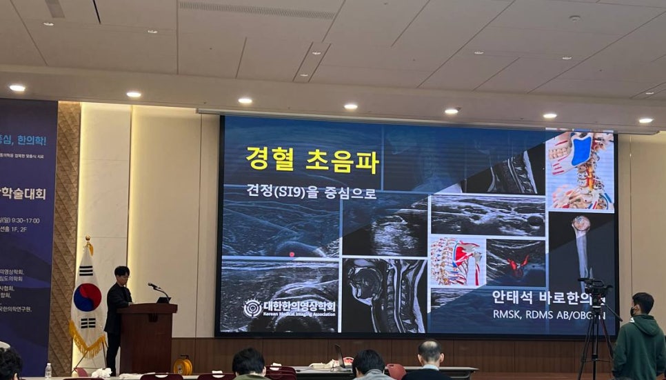 한의학 학술대회에서 강사가 경혈 초음파를 주제로 발표하고 있으며, 스크린에 견정(SI9) 부위의 초음파 영상과 해부학적 그림이 나타나 있습니다.