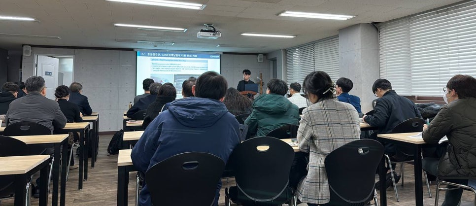 강의실에서 여러 명이 앉아 스크린의 발표 자료를 보며 강사의 설명을 듣고 있다.