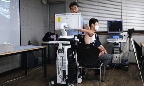 강사가 앉아있는 남성의 어깨에 초음파 기기를 대고 시연하며, 옆 모니터에 초음파 영상이 보인다.