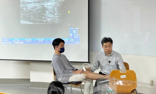 강사가 한 사람의 다리에 초음파 기기를 사용하여 시연하고 있으며, 뒤쪽 화면에 초음파 영상이 보인다.