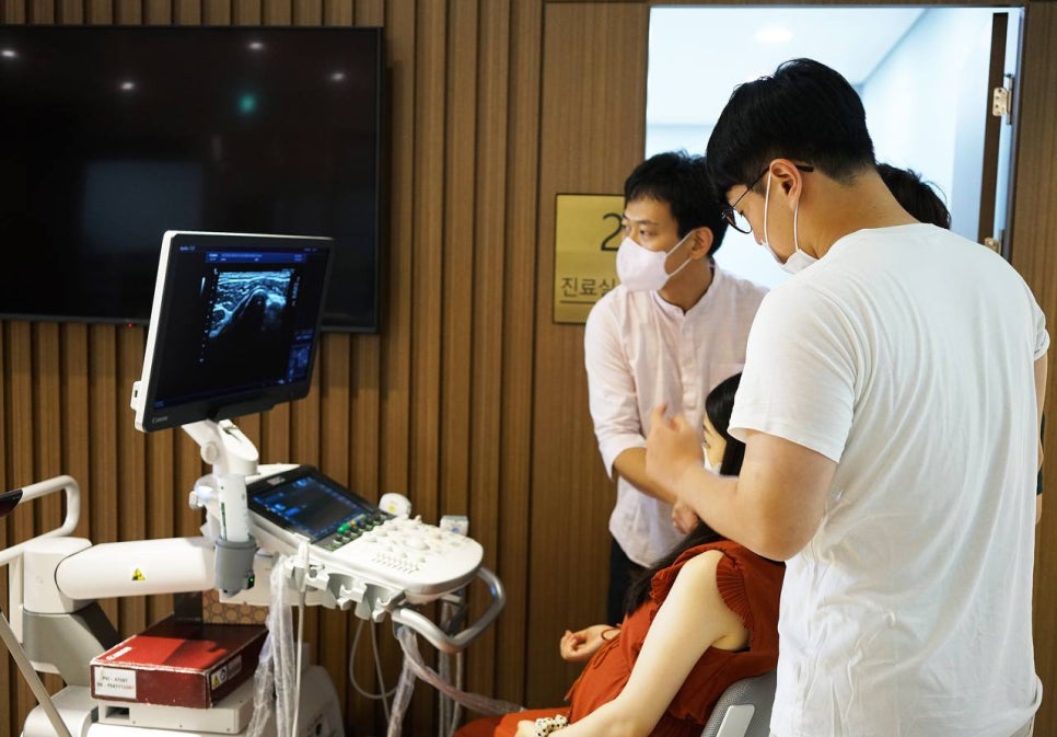 한의원 진료실에서 두 남성이 여성의 팔에 초음파 기기를 사용하며 화면을 보고 있는 모습