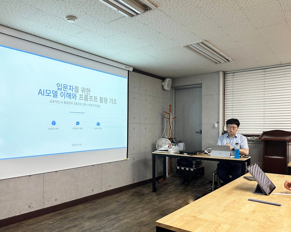 강의실에서 한 남성이 노트북 앞에 앉아 카메라를 응시하고 있으며, 뒤편 스크린에는 'AI 모델 이해와 프롬프트 활용 기초'라는 제목의 발표 자료가 투사되어 있다.