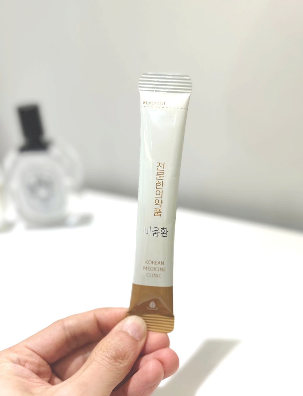 손이 '비움환'이라고 쓰인 흰색과 갈색 스틱형 한약 파우치를 들고 있는 모습.