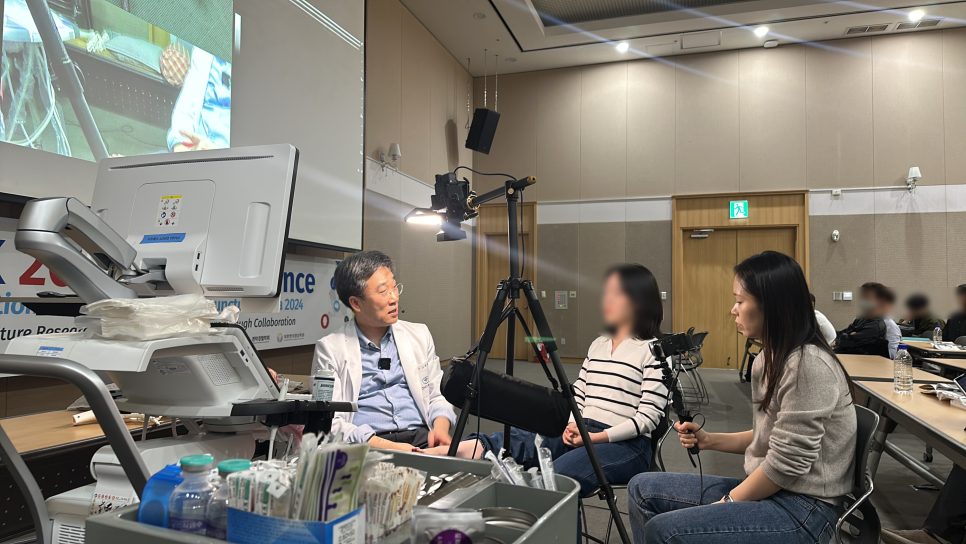한의사가 초음파 기기 옆에서 인터뷰하고 있으며, 한 여성이 촬영하고 있다.