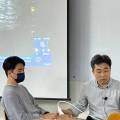 강의실에서 두 남성이 앉아 있고, 뒤쪽 스크린에 영상이 투사되고 있는 모습