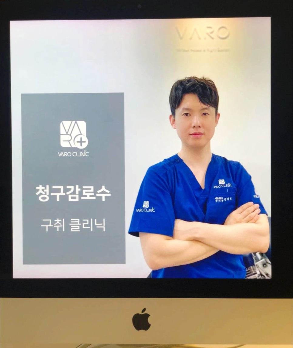 파란색 스크럽복을 입은 남성 의사가 팔짱을 끼고 모니터 앞에 서 있다. 모니터에는 'VARO CLINIC' 로고와 '청구감로수', '구취 클리닉' 문구가 보인다.