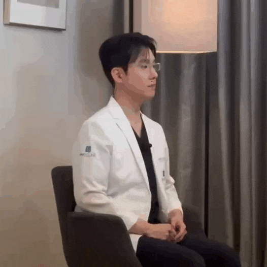흰색 의사 가운을 입고 안경을 쓴 남성이 의자에 앉아 오른쪽을 바라보고 있는 모습.