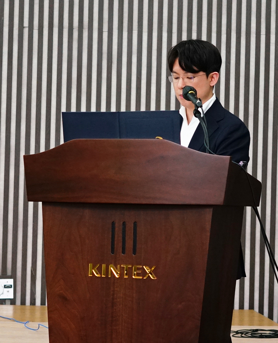 안경을 쓴 남성이 KINTEX 로고가 있는 나무 연단에서 마이크를 잡고 발표하고 있다.