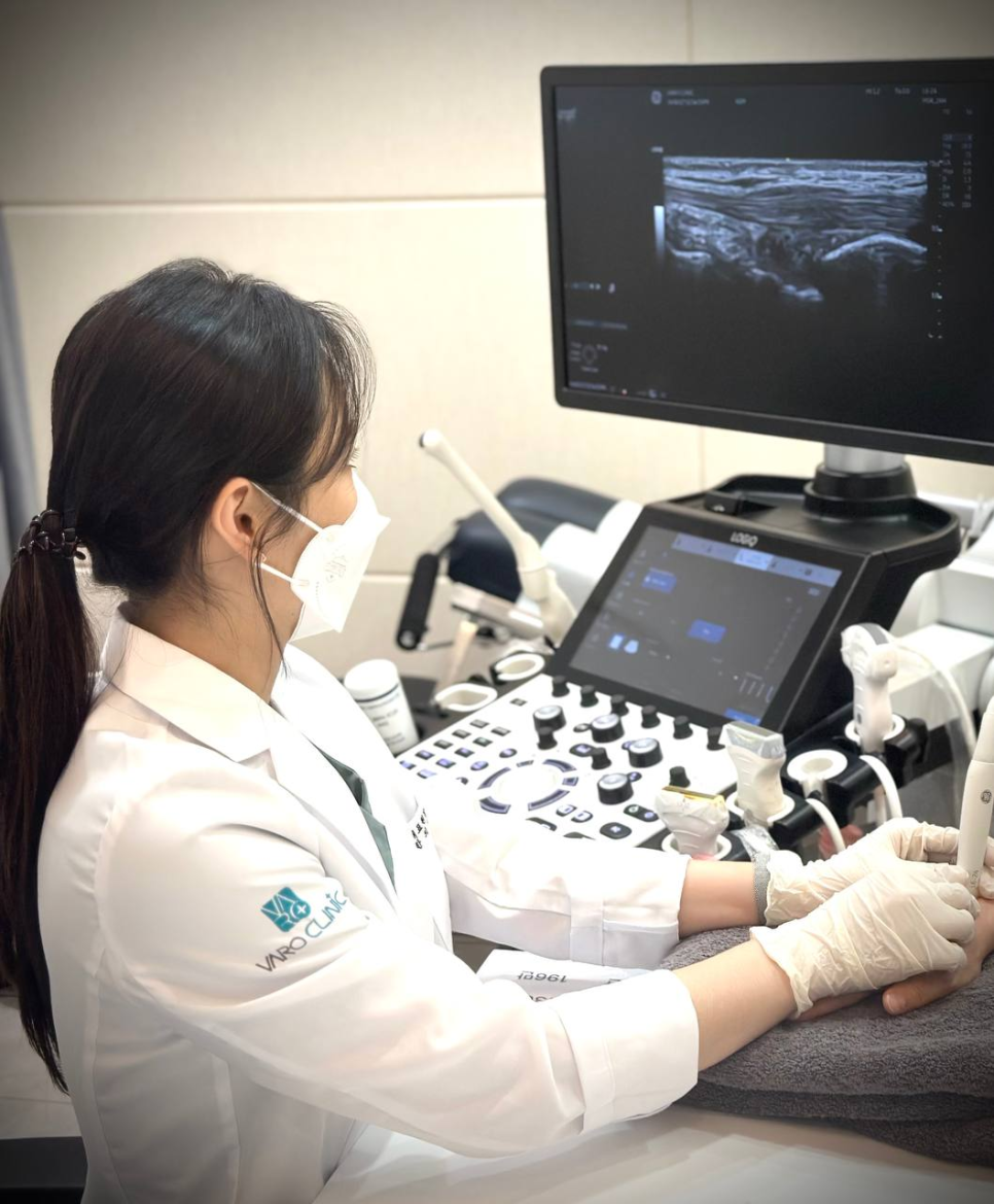 마스크와 장갑을 착용한 의료진이 환자의 손에 초음파 검사를 하고 있으며, 모니터에 초음파 영상이 보인다.