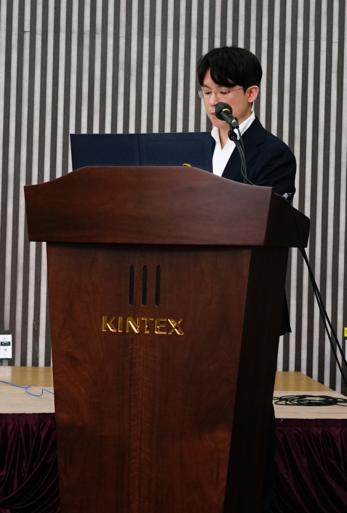 한 남성이 KINTEX 로고가 있는 나무 연단에서 마이크를 잡고 발표하고 있다.