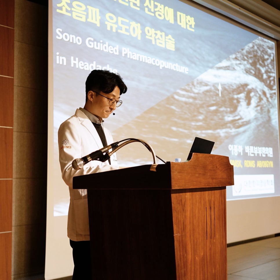 흰색 의사 가운을 입은 남성이 강단에서 발표하고 있으며, 뒤 스크린에는 초음파 유도하 약침술 관련 내용과 초음파 이미지가 표시되어 있다.