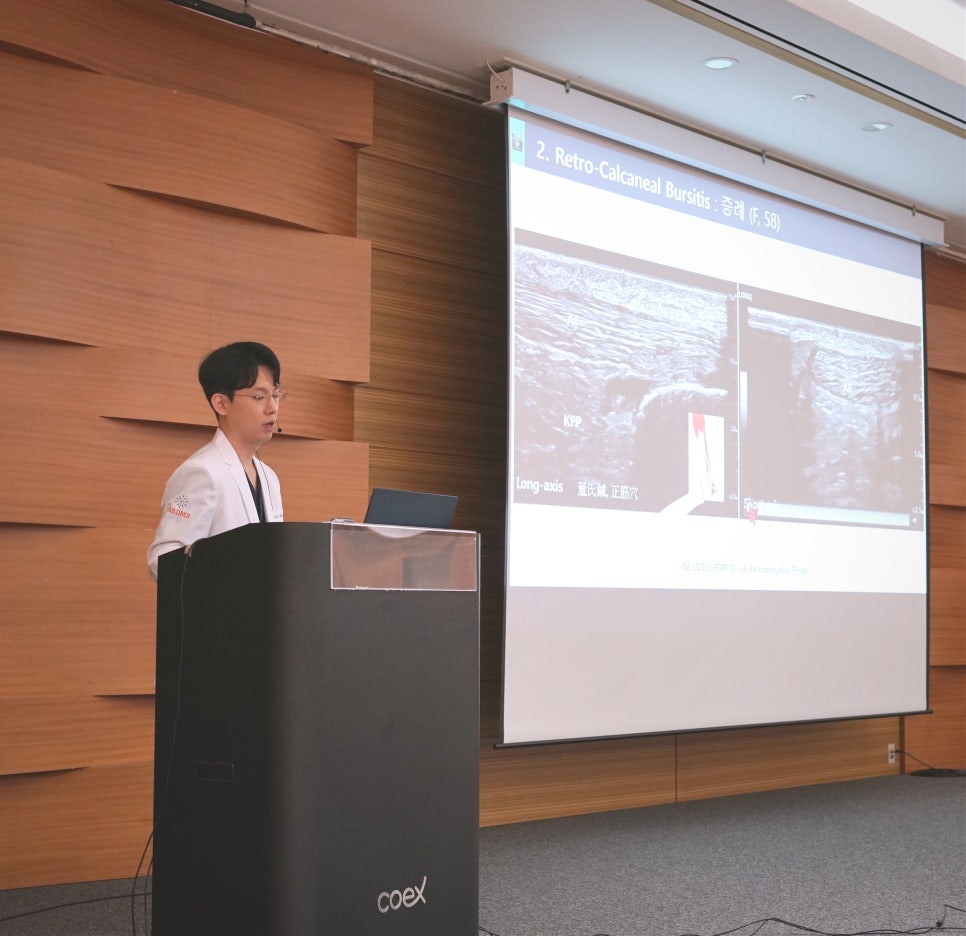 흰색 가운을 입은 남성이 연단에서 발표하고 있으며, 뒤 스크린에는 발목 초음파 영상이 보인다.