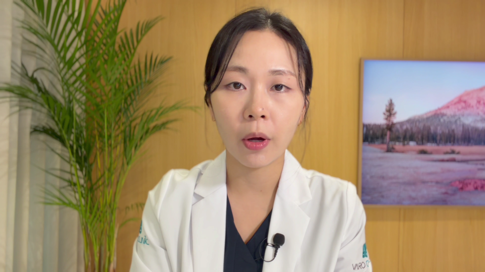 흰색 의사 가운을 입은 여성이 카메라를 보고 말하고 있으며, 배경에는 식물과 액자가 보인다.