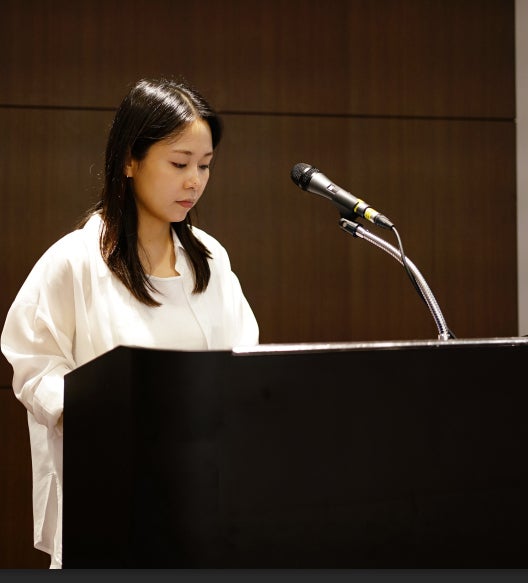 흰색 셔츠를 입은 여성이 마이크가 놓인 검은색 연단 뒤에 서서 아래를 보고 있다.