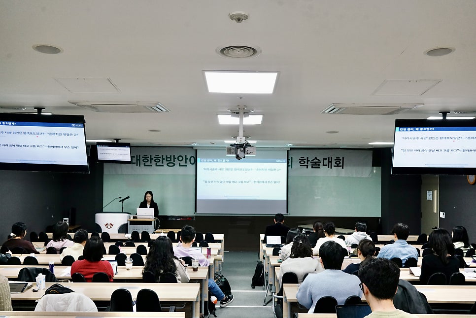 강당에서 한 여성이 발표하고 있으며, 여러 명의 참석자들이 앉아 경청하고 있다.