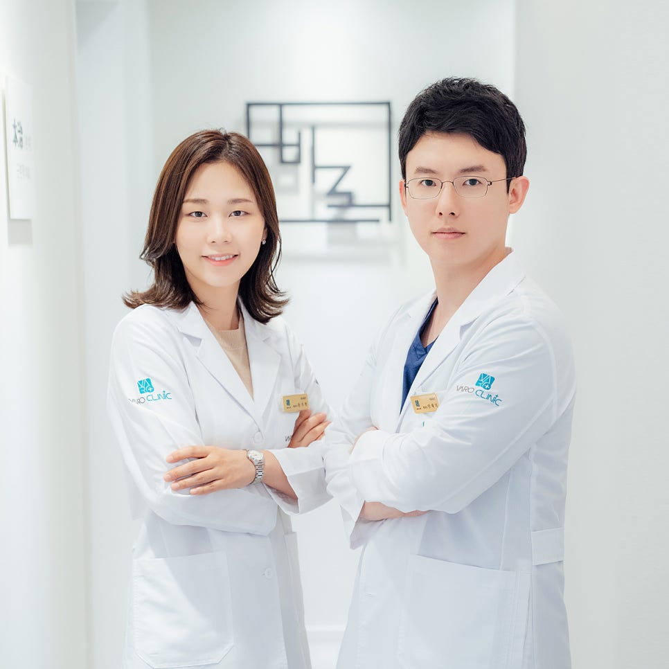 흰색 가운을 입은 남녀 한의사 두 명이 팔짱을 끼고 한의원 복도에 서서 카메라를 보고 있다.