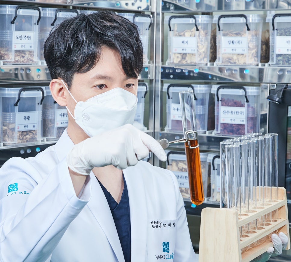 마스크와 장갑을 착용한 한의사가 흰색 가운을 입고 시험관에 담긴 주황색 액체를 집게로 들고 살펴보며, 뒤편에는 한약재가 담긴 용기들이 진열되어 있다.