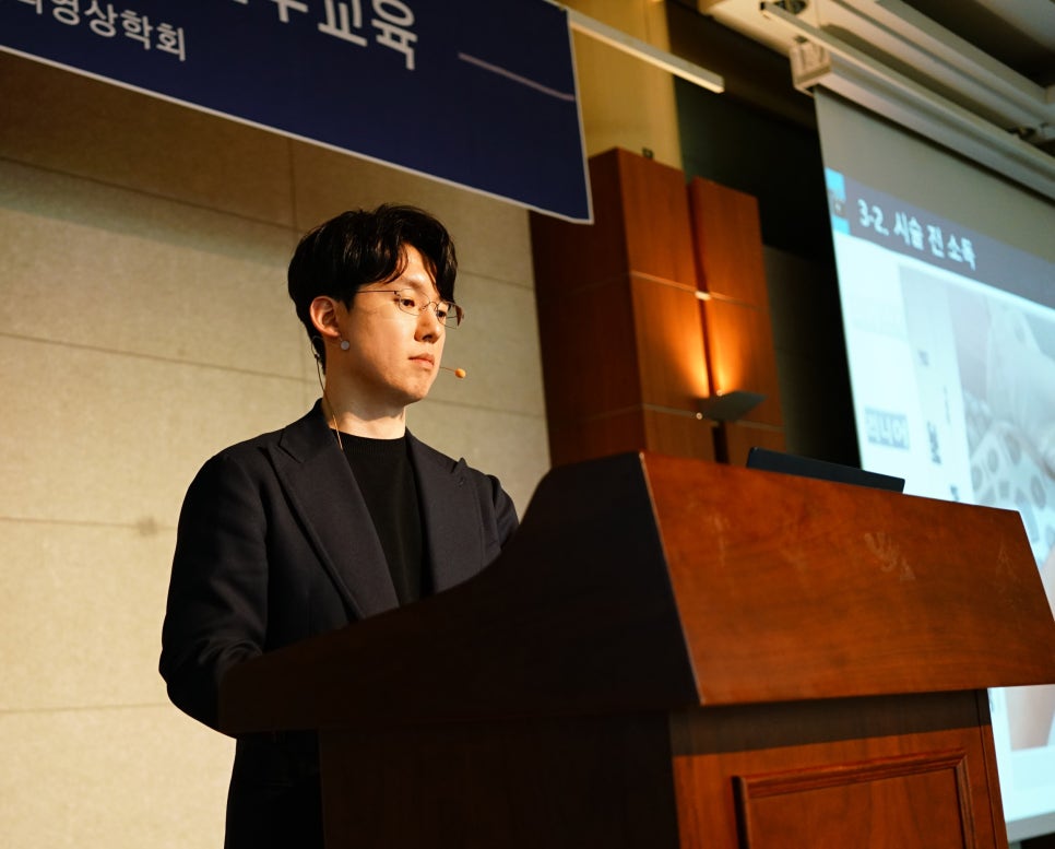 강단에서 마이크를 착용한 안경 쓴 남성이 발표하는 모습