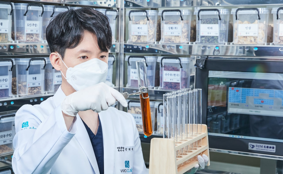 마스크를 쓴 한의사가 약재 선반 앞에서 시험관 속 액체를 관찰하고 있다 — 서울 바로한의원 초음파 약침·도침