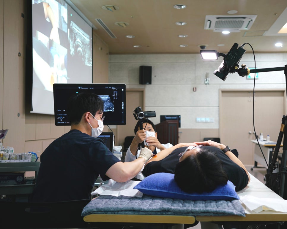 한의사가 초음파 모니터를 보며 환자에게 시술하고 있으며, 다른 한 명이 촬영하고 있는 모습.