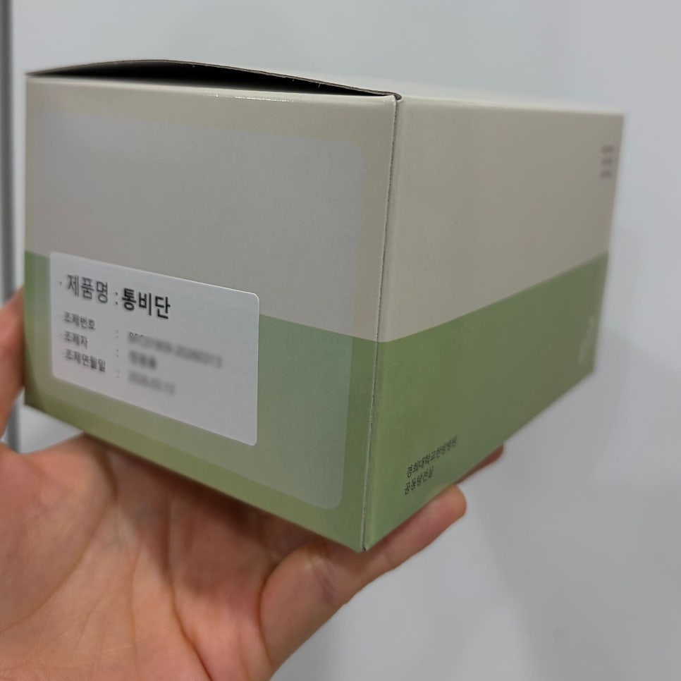 손이 '제품명: 통비단'이라고 적힌 라벨이 붙은 연두색과 베이지색 상자를 들고 있는 모습.