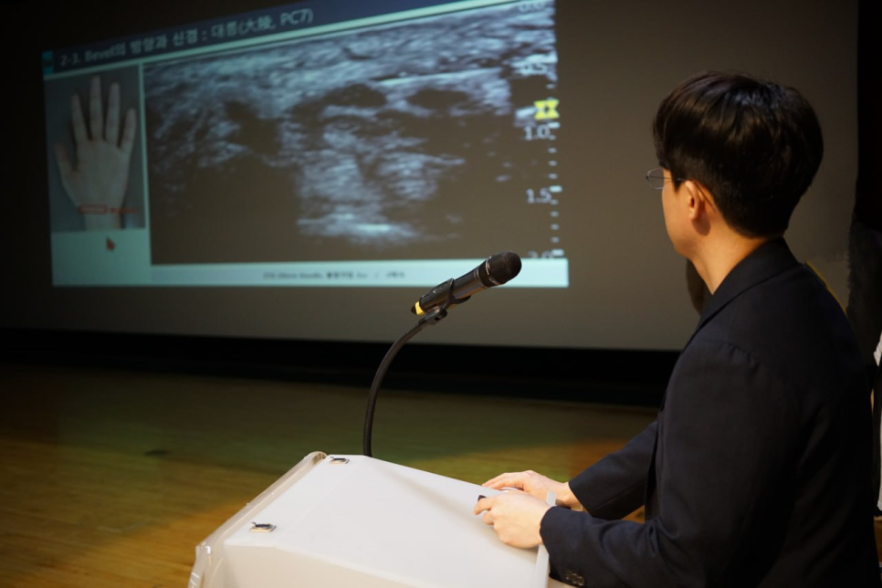 강연자가 마이크가 있는 연단에 앉아 손 그림과 초음파 영상이 보이는 스크린을 보고 있는 모습