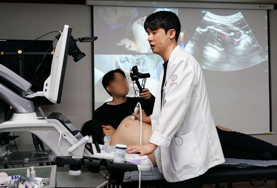 한의사가 환자에게 초음파 검사를 시연하고 있으며, 뒤편 스크린에 초음파 영상이 표시되어 있다.