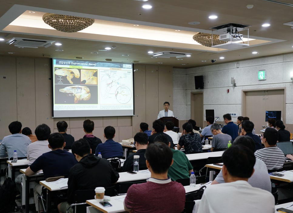 강사가 해부학 그림이 보이는 스크린 앞에서 강연하고 있으며, 여러 사람이 앉아 경청하는 모습