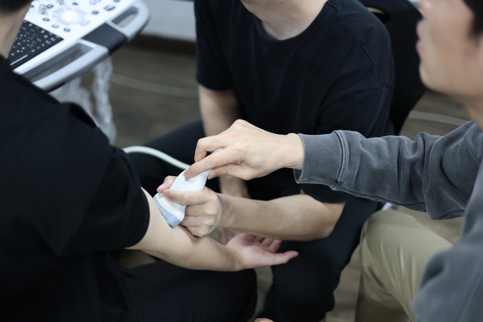 두 사람이 한 사람의 팔에 초음파 탐촉자를 대고 실습하는 모습