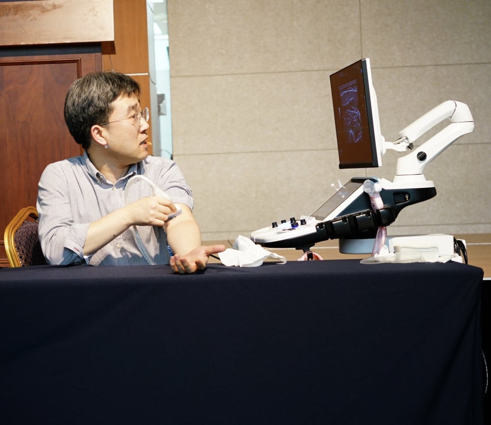 한 남성이 팔에 초음파 기기를 대고 있으며, 옆에 있는 초음파 기기 화면에 영상이 보인다.