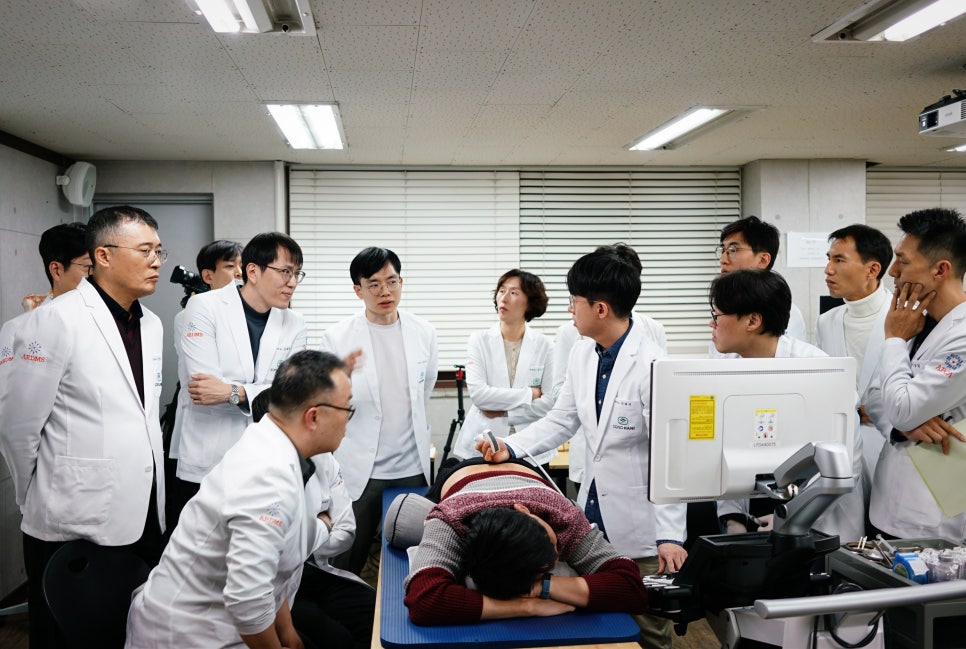 여러 명의 한의사들이 침대에 엎드린 환자에게 초음파 탐촉자를 대고 모니터를 보며 실습하는 모습 — 초음파 유도하 약침 시술 | 서울 바로한의원