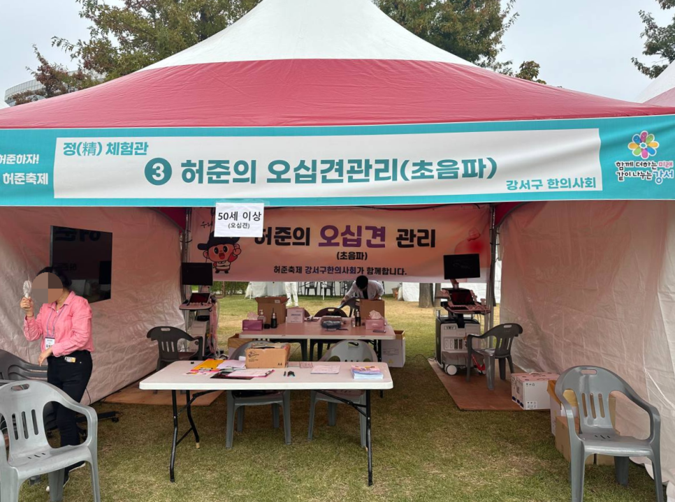 허준축제에 설치된 빨간색과 흰색 천막 부스 내부 모습으로, 테이블과 의자, 초음파 기기가 놓여 있고 한 사람이 서 있다.