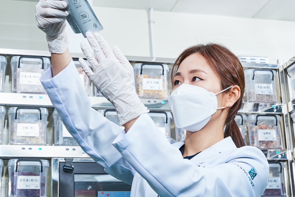 흰색 가운과 마스크를 착용한 사람이 장갑을 낀 손으로 한약 파우치를 들고 약재 선반 앞에서 확인하고 있다.