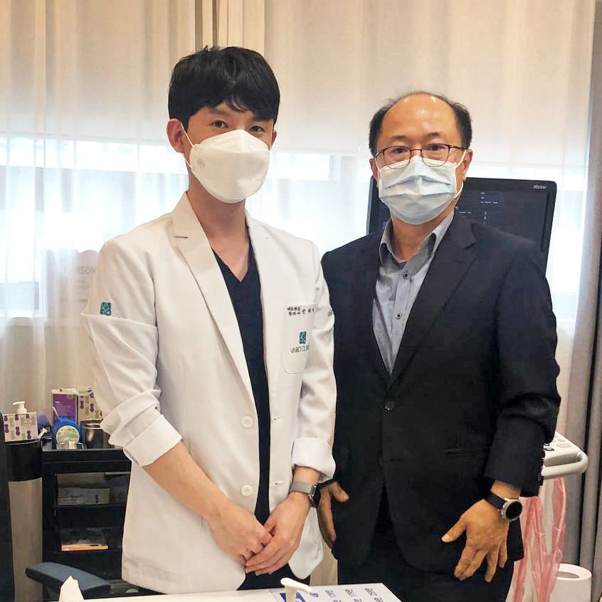 마스크를 착용한 두 남성이 나란히 서 있다. 한 명은 흰색 의사 가운을, 다른 한 명은 정장을 입고 있다.