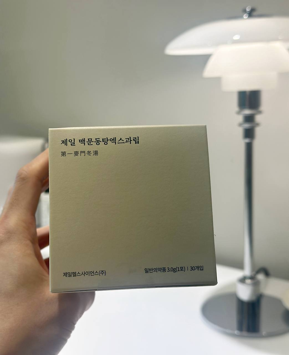 손이 '제일 맥문동탕엑스과립'이라고 적힌 연갈색 상자를 들고 있고, 배경에는 흰색 스탠드 조명이 보인다.