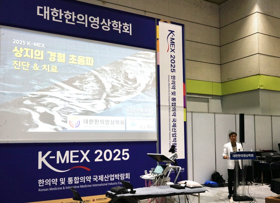 한 남성이 '대한한의영상의학회'와 '상지의 경혈 초음파 진단 & 치료'가 적힌 화면 앞에서 강연하고 있으며, 초음파 기기와 침대가 놓여 있다.