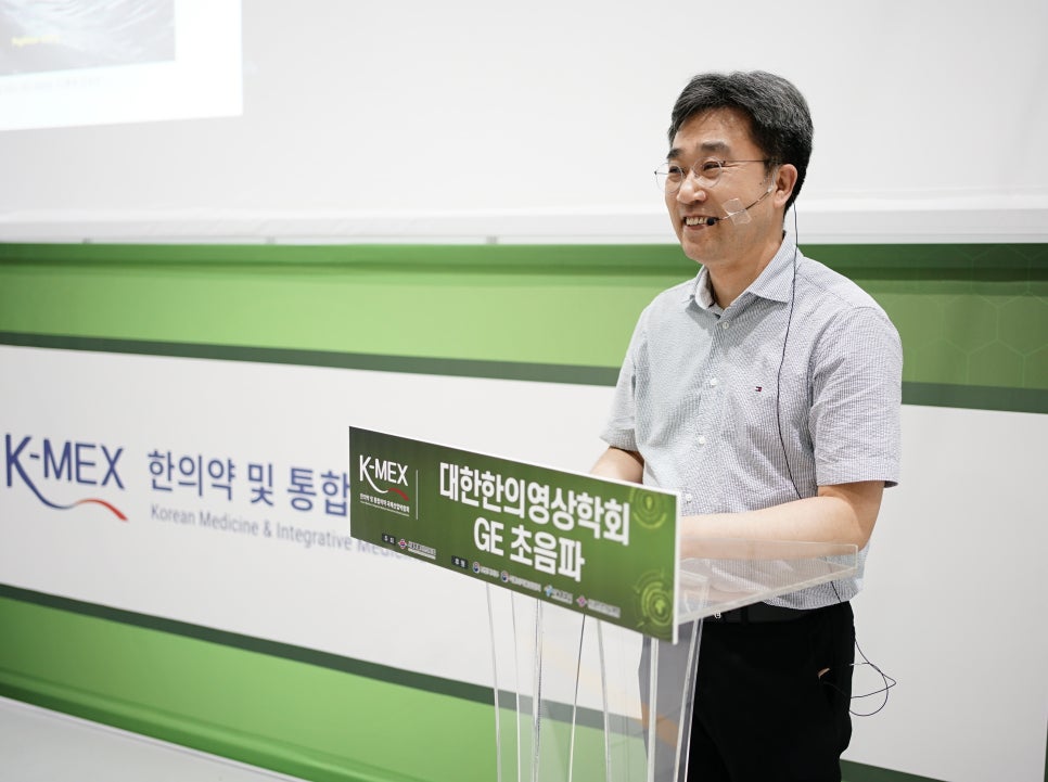 안경을 쓴 남성이 마이크를 착용하고 연단 뒤에서 웃으며 강연하고 있다. 연단에는 '대한한의영상학회 GE 초음파'라고 적힌 녹색 표지판이 있다. 배경에는 'K-MEX 한의학 및 통합' 로고가 보인다.